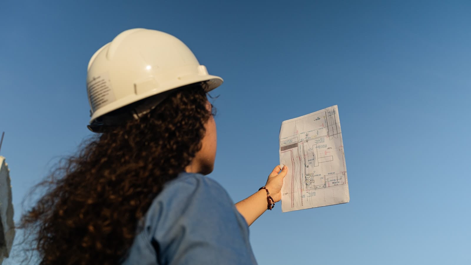 شخص يرتدي خوذة سلامة بيضاء يحمل مخططاً هندسياً في موقع عمل خارجي