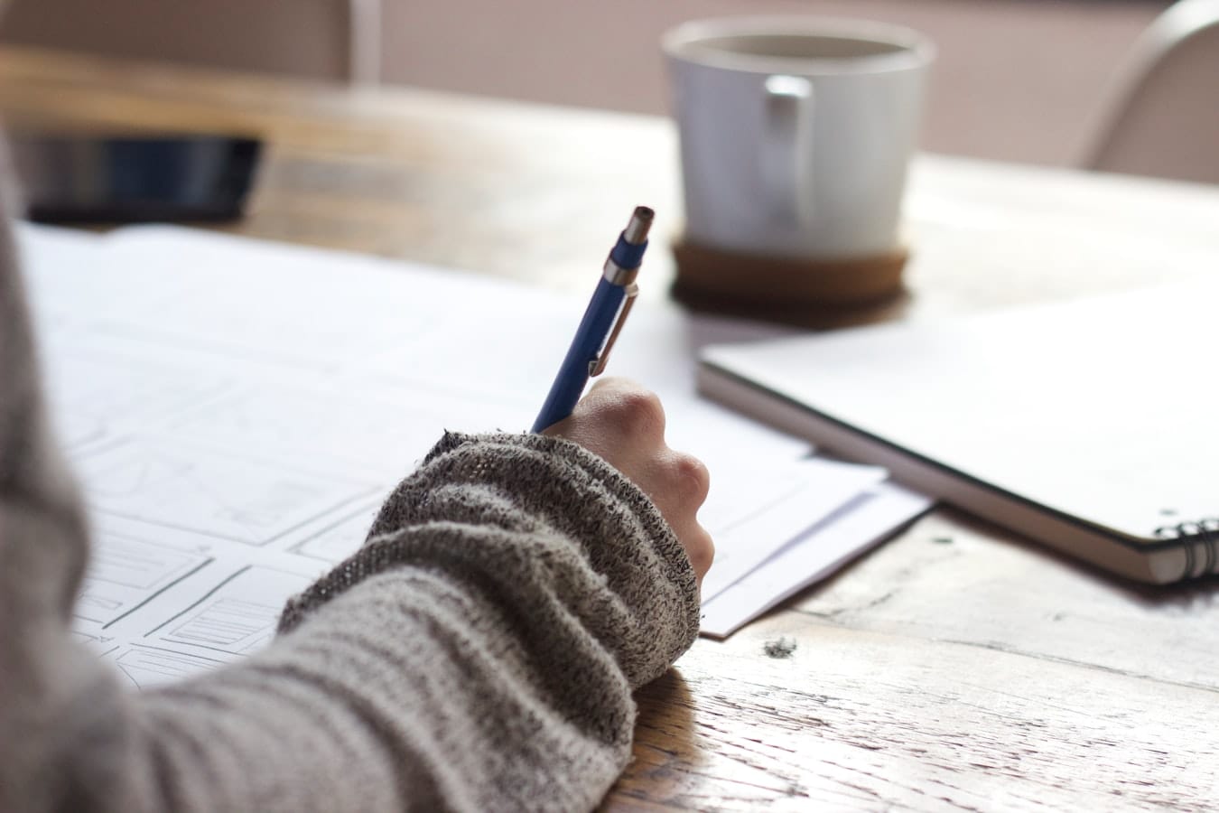 Person in a gray sweater sketching wireframe diagrams on paper at a wooden table